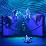 A group of dancers on a stage in front of a blue curtain.